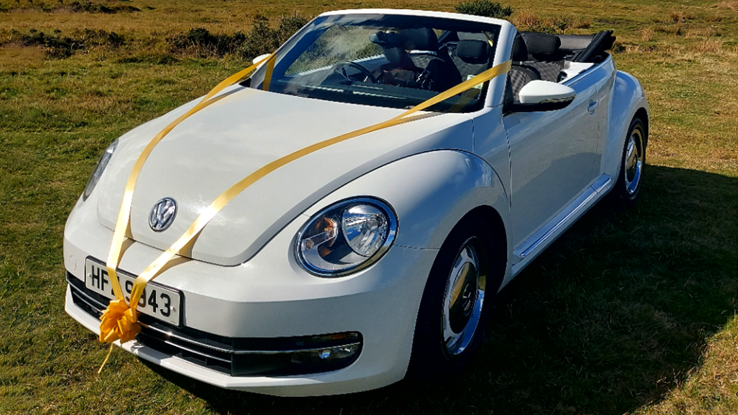 Front view of a white Volkswagen Beetle with convertible open decorated with gold wedding ribbons, parked outdoors on grass with clear skies above.