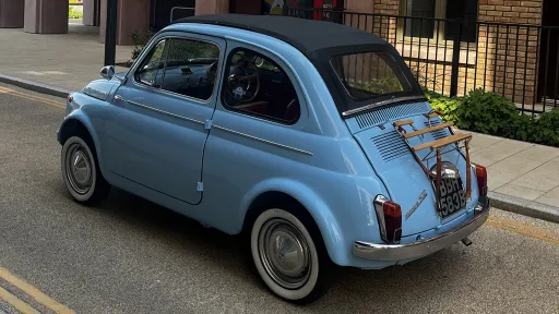 Left rear side view of a blue Fiat 500 convertible with whitewall tyres, parked on a city street beside brick buildings.