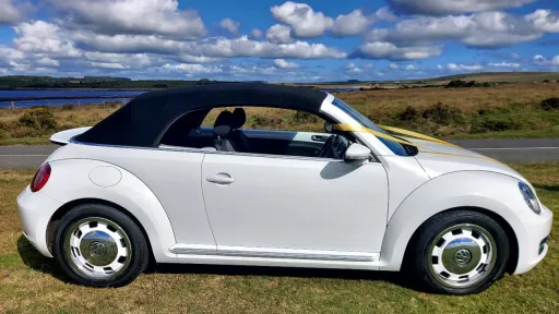 Right side view of a white Volkswagen Beetle convertible with contrasting black roof, parked beside a country road with open fields in the background.