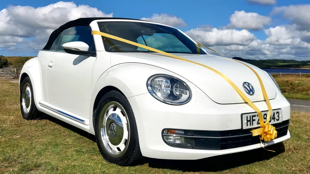 White modern Volkswagen Beetle convertible with black roof, parked on grass in Devon countryside under a blue sky.