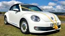 White modern Volkswagen Beetle convertible with black roof, parked on grass in Devon countryside under a blue sky.