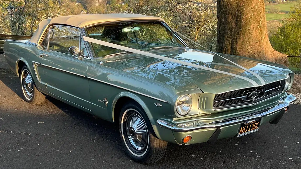 Front angled view of a green classic Ford Mustang convertible with white soft-top roof closed, parked under trees on a country lane.