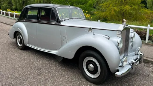Front angled view of a 1954 Rolls-Royce Silver Dawn in silver with black roof, parked on a gravel driveway with greenery in the background.