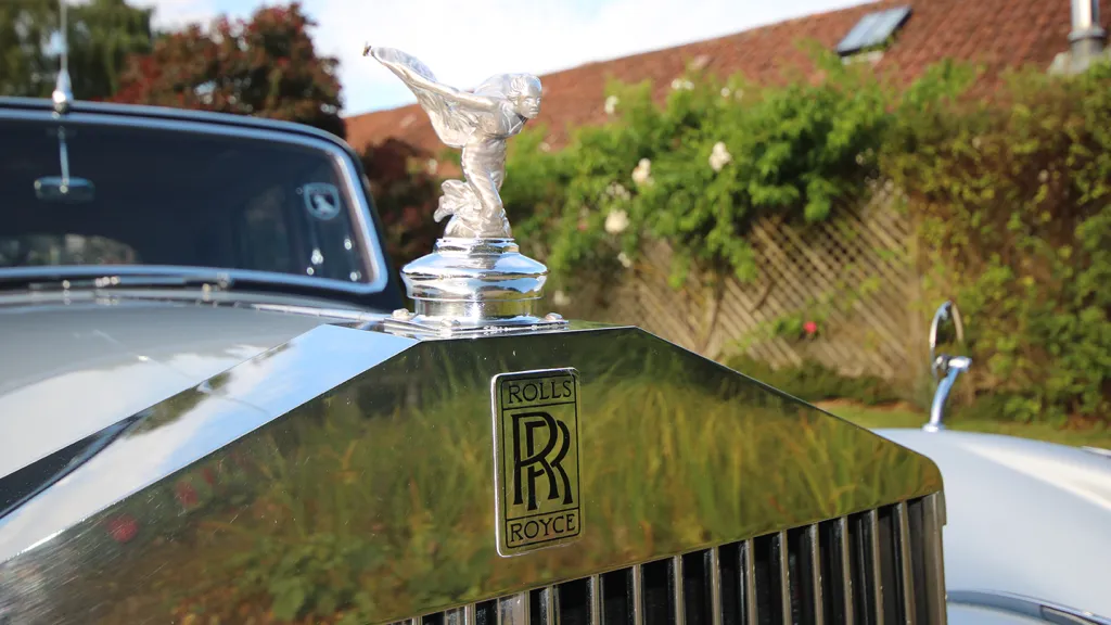 Close-up of the Rolls-Royce Spirit of Ecstasy bonnet mascot and chrome grille on a 1954 Silver Dawn with blurred garden background.