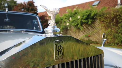 Close-up of the Rolls-Royce Spirit of Ecstasy bonnet mascot and chrome grille on a 1954 Silver Dawn with blurred garden background.