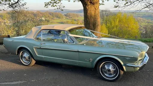 Green classic Ford Mustang convertible with white soft-top roof, parked outdoors on a country road for wedding hire.