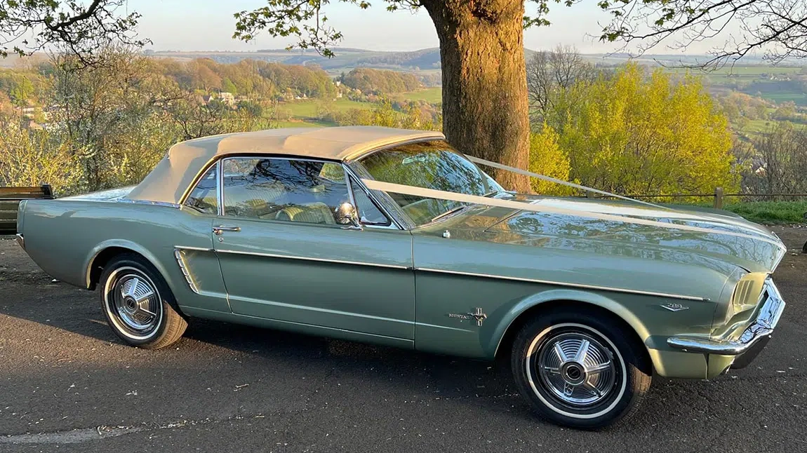 Green classic Ford Mustang convertible with white soft-top roof, parked outdoors on a country road for wedding hire.