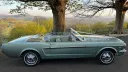Right side view of a green classic Ford Mustang convertible with roof folded down, showing open interior seating, parked in countryside setting.