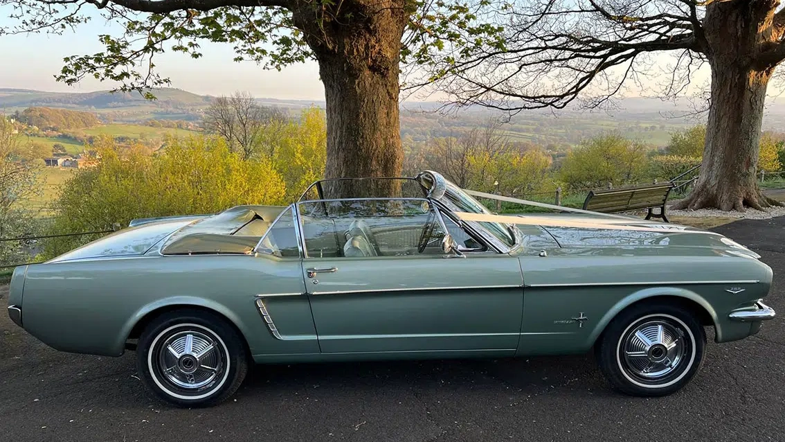 Right side view of a green classic Ford Mustang convertible with roof folded down, showing open interior seating, parked in countryside setting.