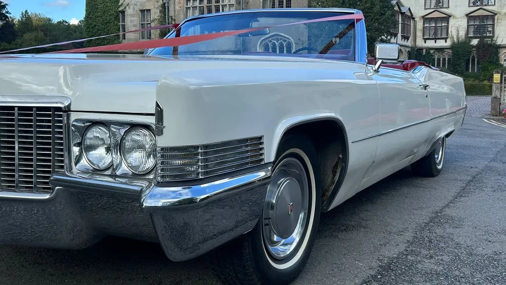 Front angled view of a 1970 white Cadillac Coupe DeVille convertible with red leather interior, parked outside a grand building.