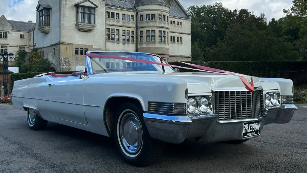White 1970 Cadillac Coupe DeVille convertible with red leather interior and wedding ribbons on the bonnet, parked outside a stately venue.