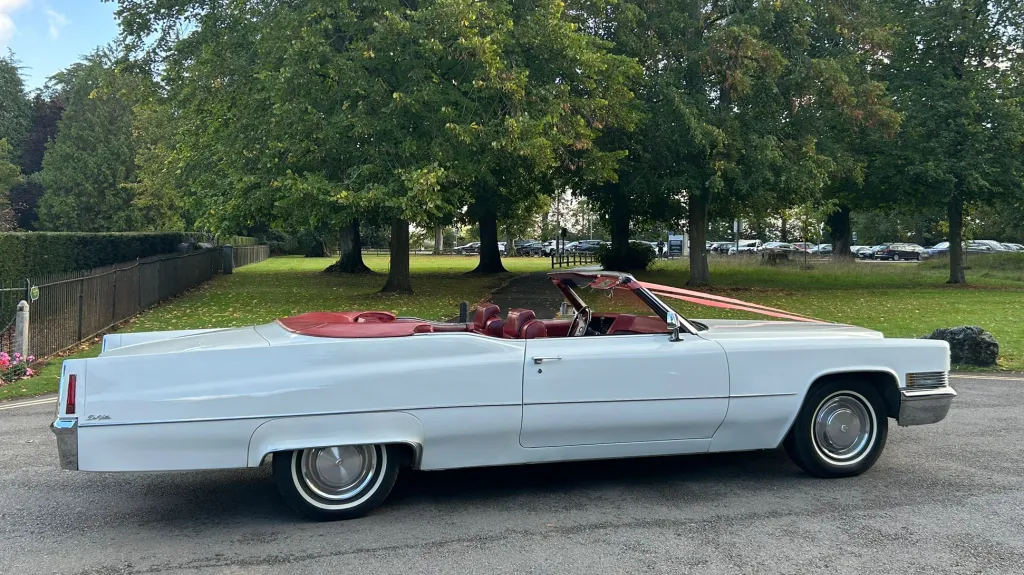 Right side profile of a white 1970 Cadillac Coupe DeVille convertible with red leather interior and roof folded down, parked on grass.