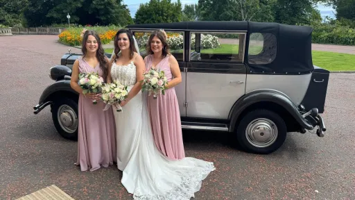 Bride and bridesmaids posing beside a silver and grey Regent convertible wedding car with white ribbons, parked on a paved driveway.
