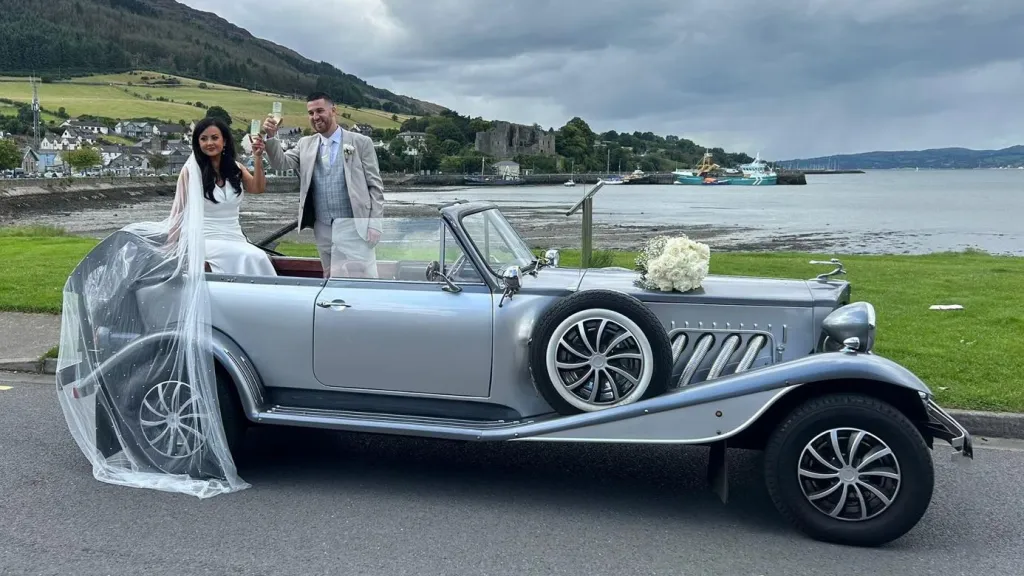 Bride and groom standing beside a silver Beauford convertible wedding car with black soft-top roof, parked near a lake close to Belfast.