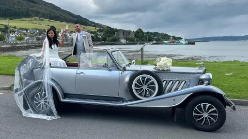 Bride and groom standing beside a silver Beauford convertible wedding car with black soft-top roof, parked near a lake close to Belfast.