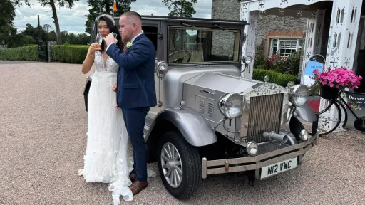 Bride and groom standing beside a silver Imperial wedding car decorated with white ribbons, parked outside a stone venue.