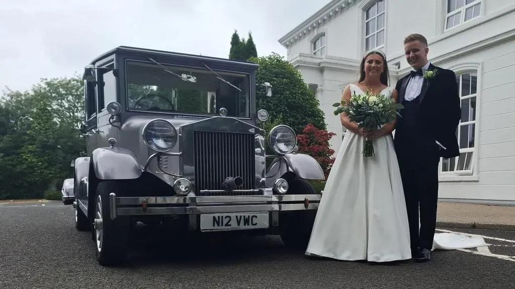 Silver and black Imperial vintage-style wedding car parked beside a white country house with the bride and groom posing beside it.