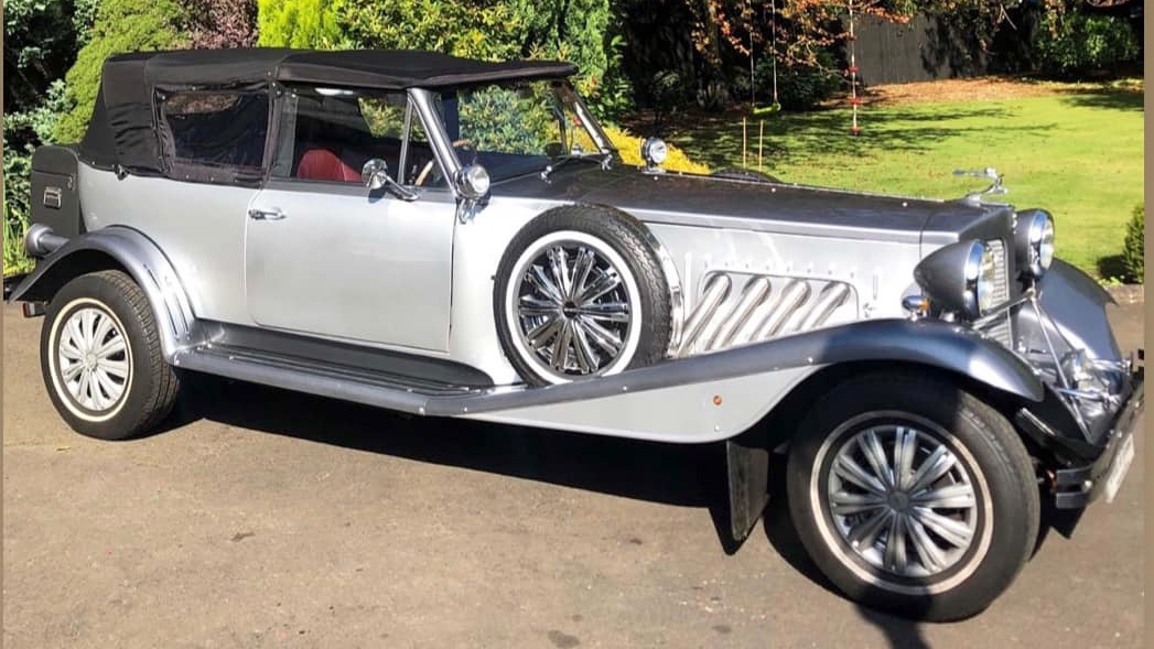 Right side profile of a silver Beauford convertible with black soft-top roof and chrome detailing, parked on a country driveway.