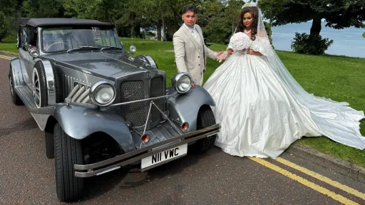 Bride and groom posing with a silver Beauford convertible decorated with white wedding ribbons, photographed in a scenic countryside setting in Ireland.