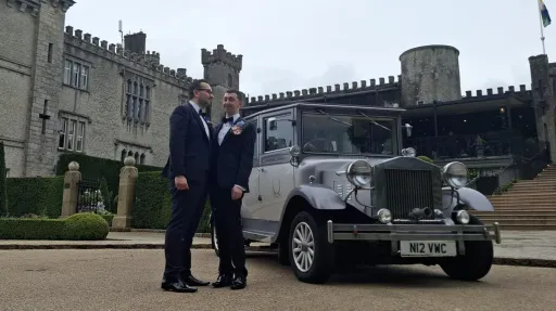 Two grooms standing beside a silver Imperial vintage wedding car outside a castle venue in Northern Ireland.