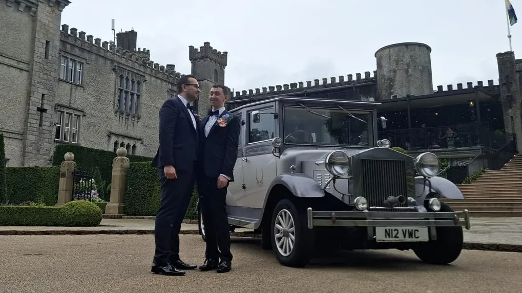 Two grooms standing beside a silver Imperial vintage wedding car outside a castle venue in Northern Ireland.