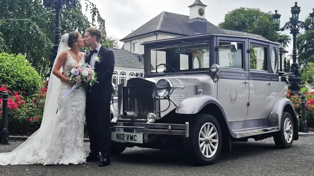 Silver Imperial vintage wedding car with white ribbons parked on a driveway surrounded by greenery.