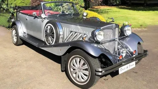 Front angled view of a silver and grey Beauford vintage-style convertible wedding car with chrome headlights and spare side wheels, parked on a sunny driveway.