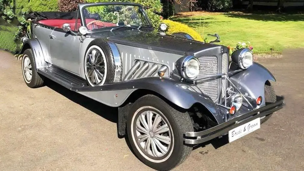 Front angled view of a silver and grey Beauford vintage-style convertible wedding car with chrome headlights and spare side wheels, parked on a sunny driveway.