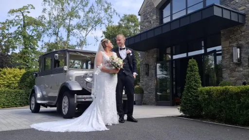 Bride and groom posing beside a silver Imperial wedding car in front of a modern hotel building.