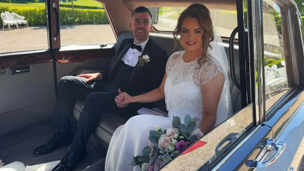 Bride and groom seated inside a silver Daimler limousine with grey leather interior, smiling and holding the bridal bouquet.
