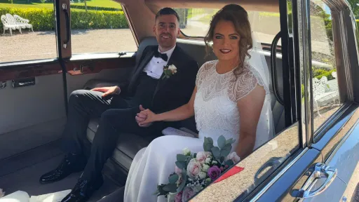Bride and groom seated inside a silver Daimler limousine with grey leather interior, smiling and holding the bridal bouquet.