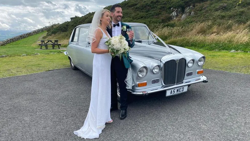 Bride and groom posing beside a silver Daimler DS420 limousine decorated with white ribbons, parked on a country road.