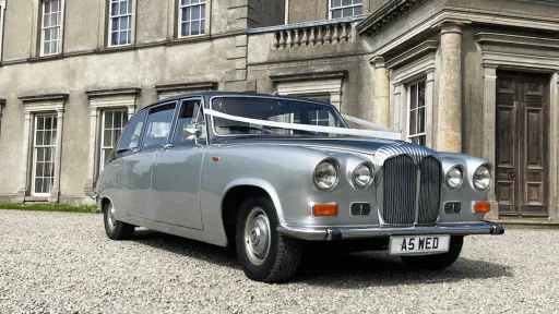 Front angled view of a silver Daimler DS420 limousine with chrome grille and twin headlights, parked outside a stately building.