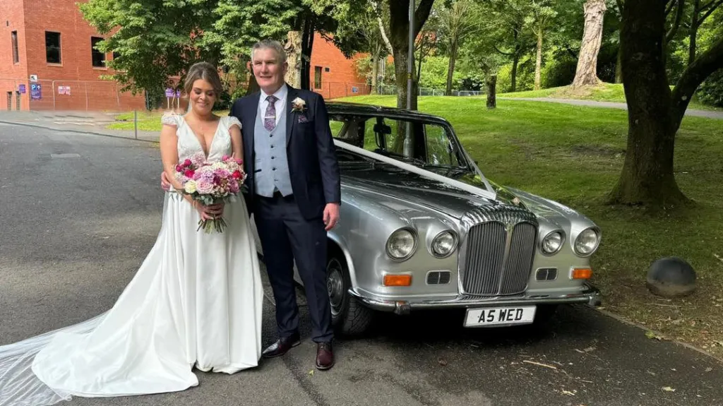 Bride and groom posing next to a silver Daimler DS420 limousine parked on a tree-lined driveway in Antrim.