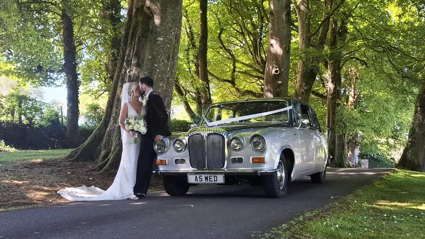 Bride and groom photographed beside a silver Daimler limousine with chrome details, parked under tall trees in Belfast.