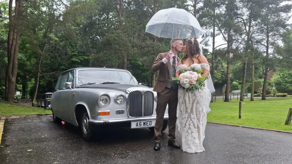 Happy couple walking under an umbrella beside a silver Daimler limousine on a rainy wedding day.