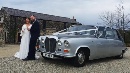 Bride and groom beside a silver Daimler DS420 limousine, parked in front of a rustic barn venue in Northern Ireland.