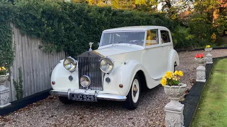 Side view of a 1948 white Rolls-Royce Silver Wraith parked on a gravel driveway, featuring chrome headlights and a classic Rolls-Royce grille.