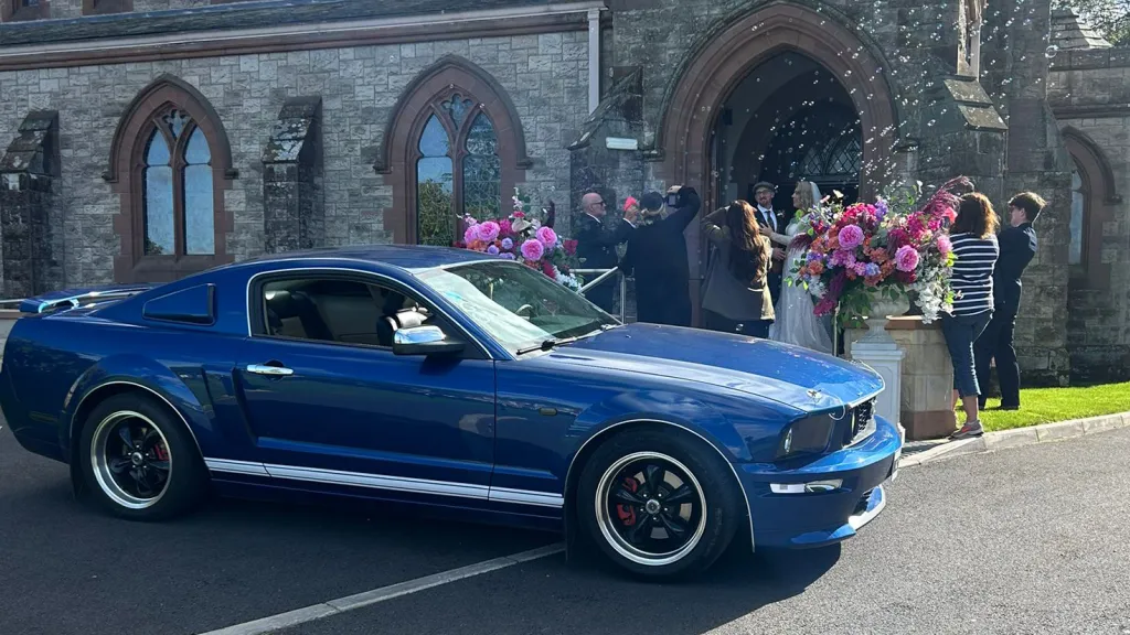 Blue Ford Mustang parked outside a church in Antrim, Northern Ireland, decorated with white ribbons for a wedding ceremony.