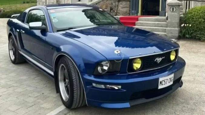 Front angled view of a blue Ford Mustang convertible with wedding ribbons, parked on a gravel driveway beside a church.