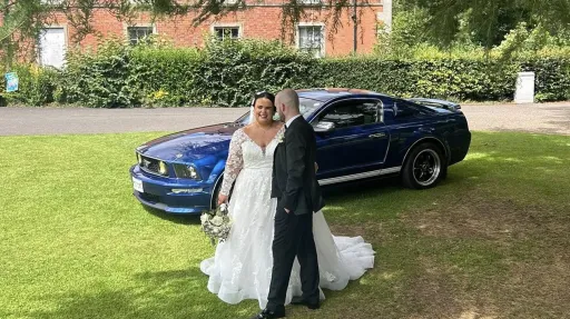 Bride and groom standing beside a blue Ford Mustang wedding car parked on the grass in front of a country house.