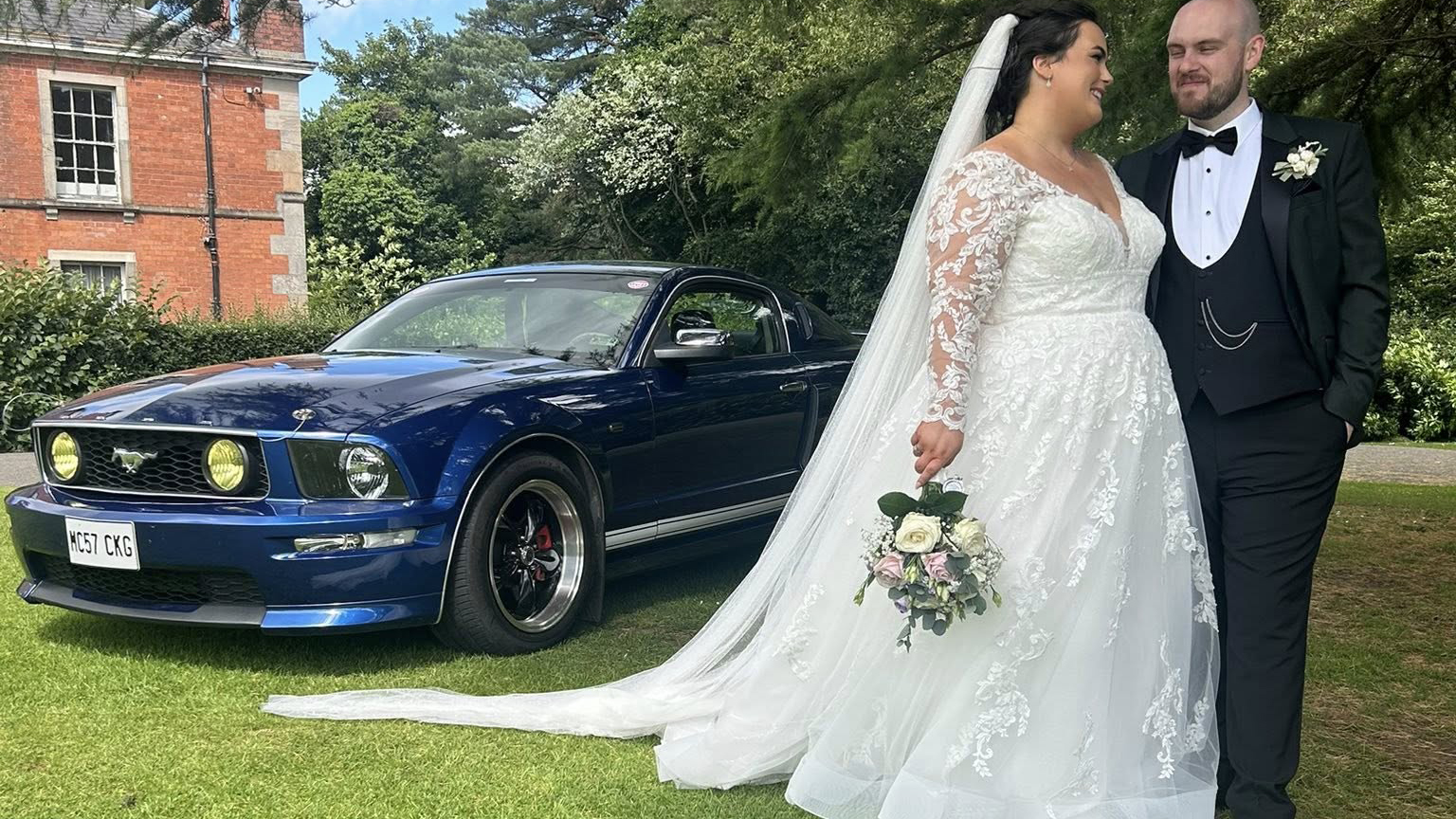 Bride and groom smiling beside a blue Ford Mustang wedding car on a sunny day, surrounded by greenery.