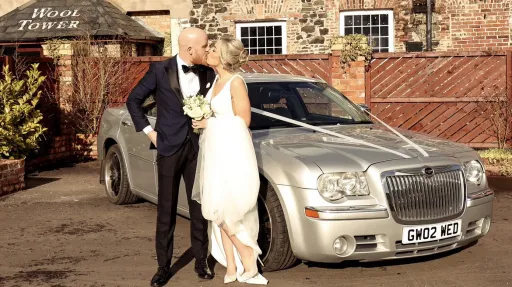 Bride and groom posing beside a silver Chrysler 300C wedding car decorated with white ribbons, parked outside a rustic brick venue.