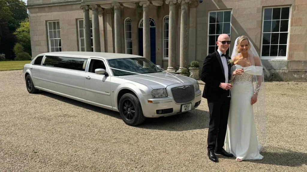 Bride and groom posing beside a silver Chrysler stretch limousine parked outside a grand country house in Northern Ireland.