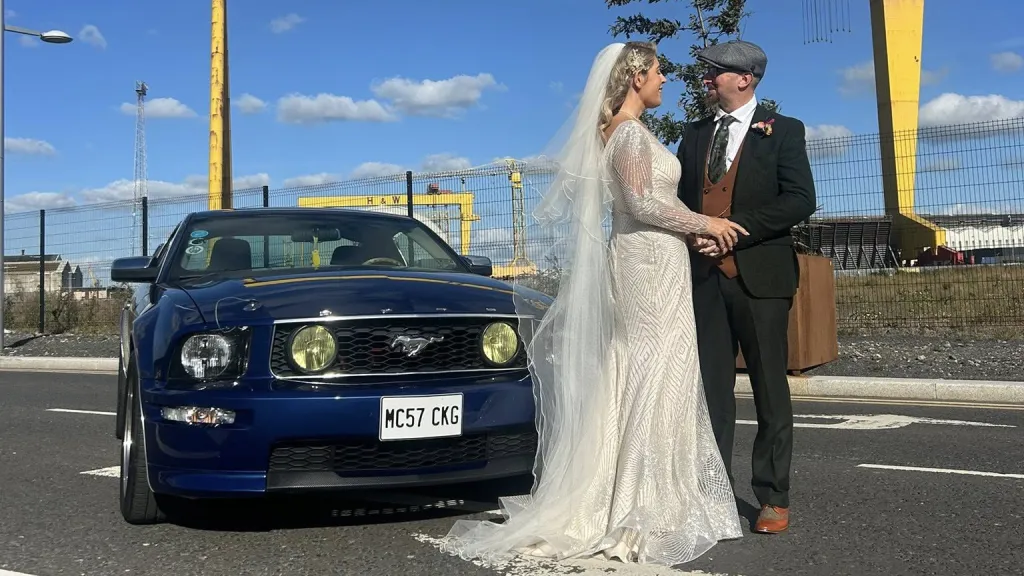 Bride and groom posing beside a blue Ford Mustang decorated with ribbons, with Northern Ireland’s Harland & Wolff cranes in the background.
