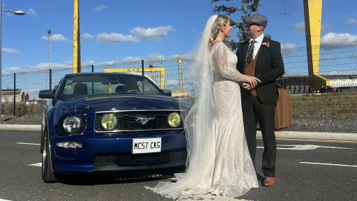 Bride and groom posing beside a blue Ford Mustang decorated with ribbons, with Northern Ireland’s Harland & Wolff cranes in the background.