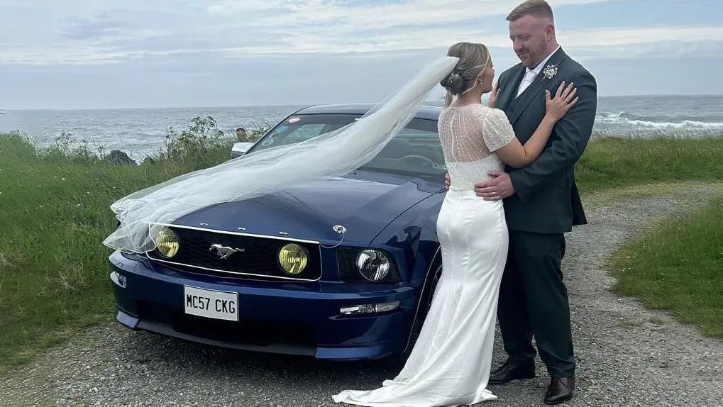 Bride and groom standing next to a blue Ford Mustang wedding car parked on a coastal road, with ribbons on the bonnet.