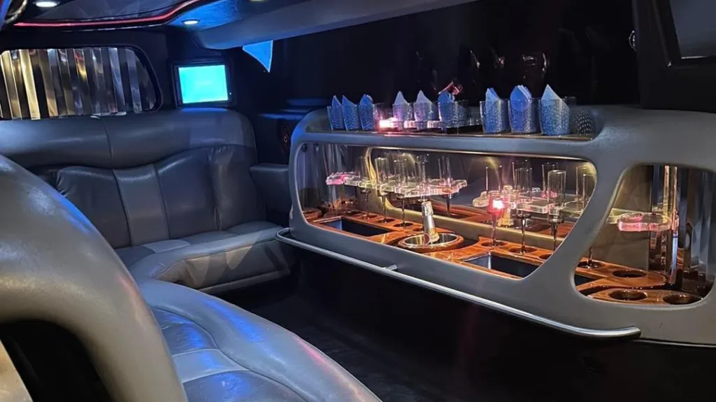 Interior of a silver Chrysler stretch limousine featuring two tone silver leather seating, neon lighting, and a built-in drinks bar with illuminated bottles.