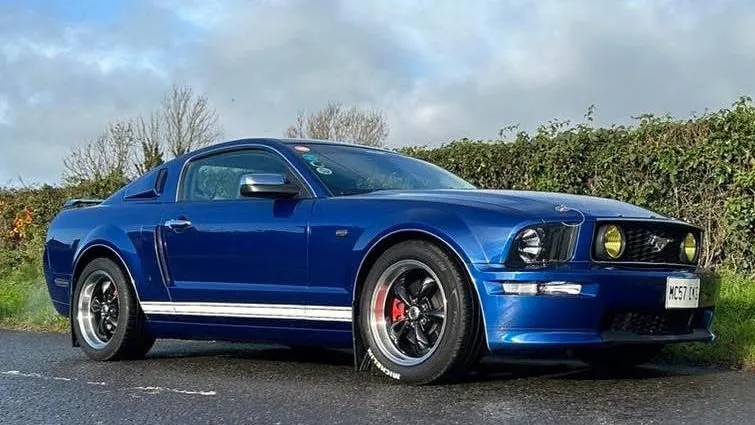 Side view of a modern blue Ford Mustang with alloy wheels, parked on a quiet countryside lane.