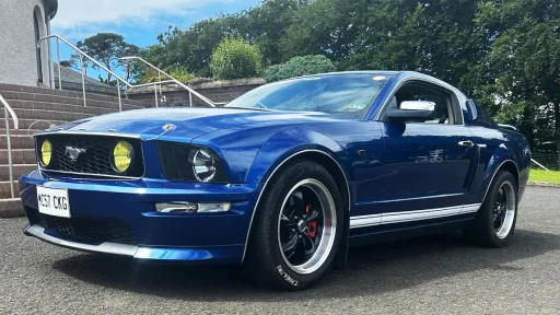 Blue Ford Mustang wedding car parked on a gravel driveway with chrome wheels and bonnet ribbons.